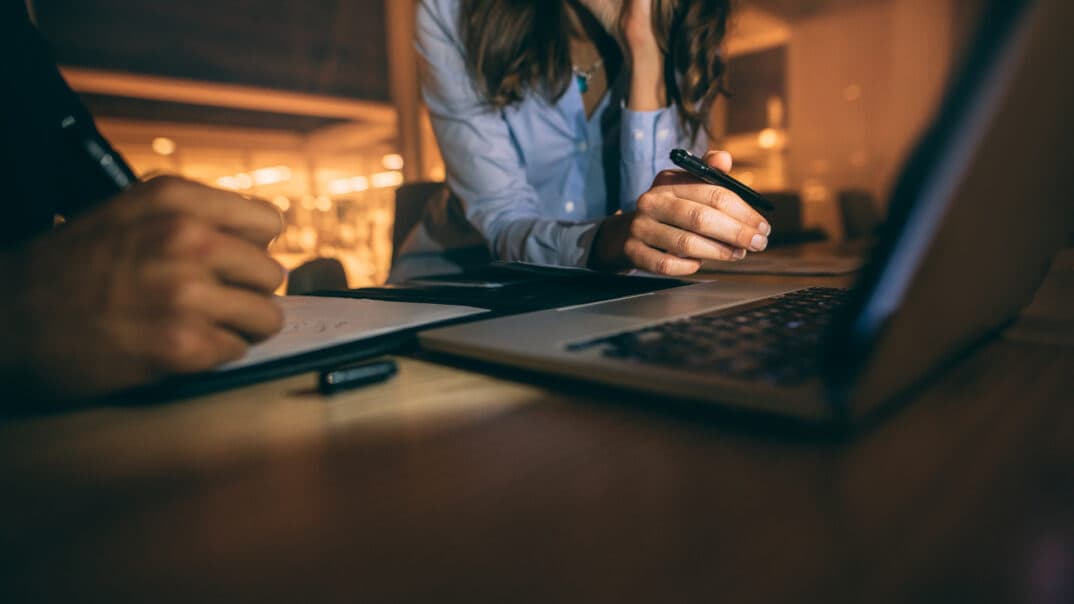 A close up of two people writing with pens in front of an open laptop